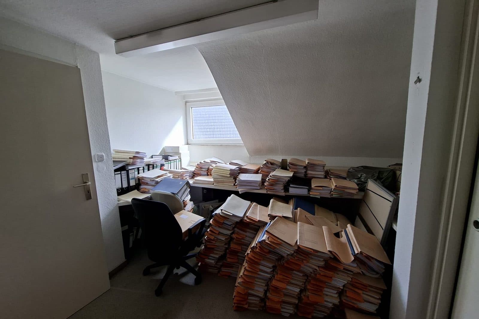 Büroraum vor der Räumung, vollgestellt mit Aktenstapeln, Schreibtisch und Bürostuhl